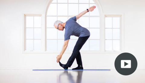 side view of tom mccook standing on mat, right leg slightly bent, left leg straight, touching left toes with left hand, right arm straight up and back; picture of play button in bottom right corner