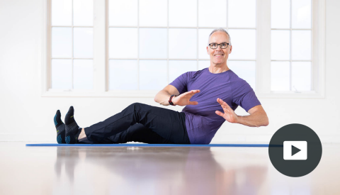 side view of tom mccook sitting on mat with legs slightly bent out in front, twisting to left side; picture of play button on bottom right corner