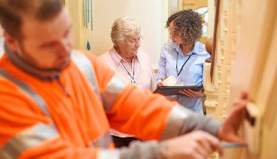 An alarm fitter fits an alarm in a senior woman's house . The tenant and housing officer watches on to make sure her tenant is happy with the installation and discuss where they will fit the smoke alarm next