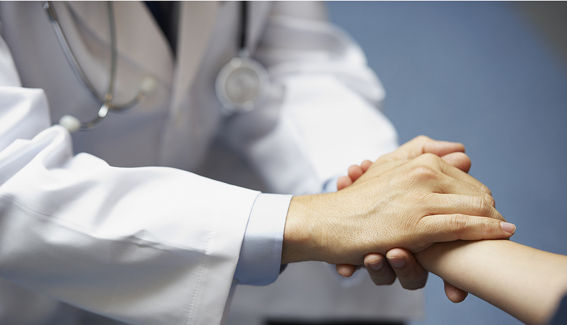 The torso of a doctor in a white lab coat with stethoscope holding a patient's hand.