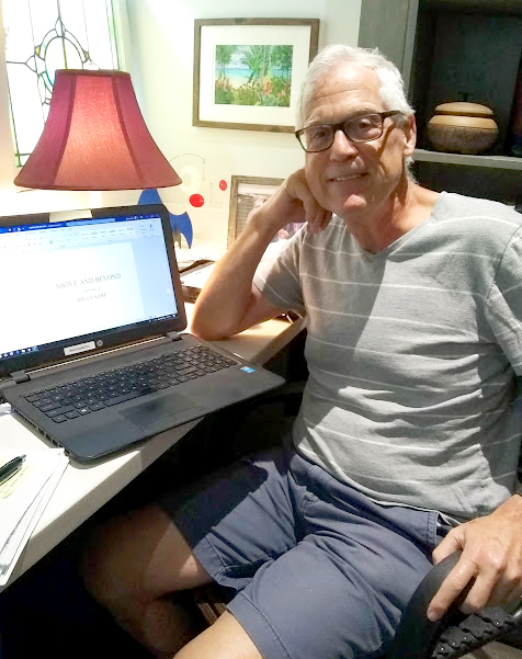 a man sitting at a desk in his home office turns from his laptop to smile for the camera and show the title of the book he is writing on the laptop monitor