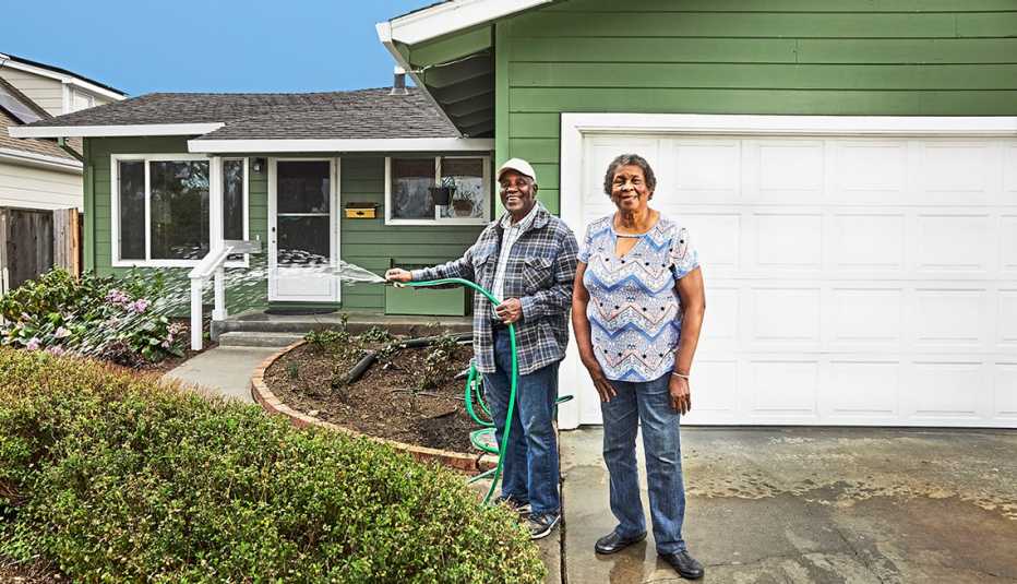 an older couple stands in the driveway of their home and the man holds a hose and is watering the front garden