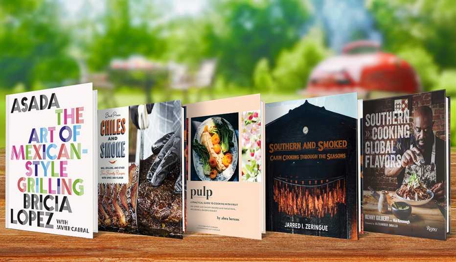 five books including asada: the art of mexican-style grilling,  chiles and smoke: b b q, grilling, and other fire-friendly recipes with spice and flavor, pulp: a practical guide to cooking with fruit, southern and smoked: cajun cooking through the seasons and southern cooking, global flavors on wooden table with blurred background of grill, table and chairs and trees
