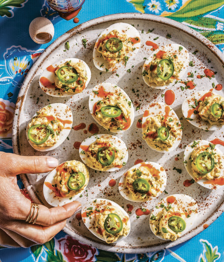 deviled eggs topped with jalapenos, hot sauce, cumin on a dish; hand grabbing one egg deviled eggs topped with jalapenos, hot sauce, cumin on a dish; hand grabbing one egg