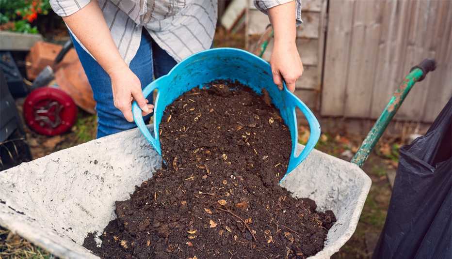 A woman tipping compost from a bucket into a wheelbarrow in her garden.