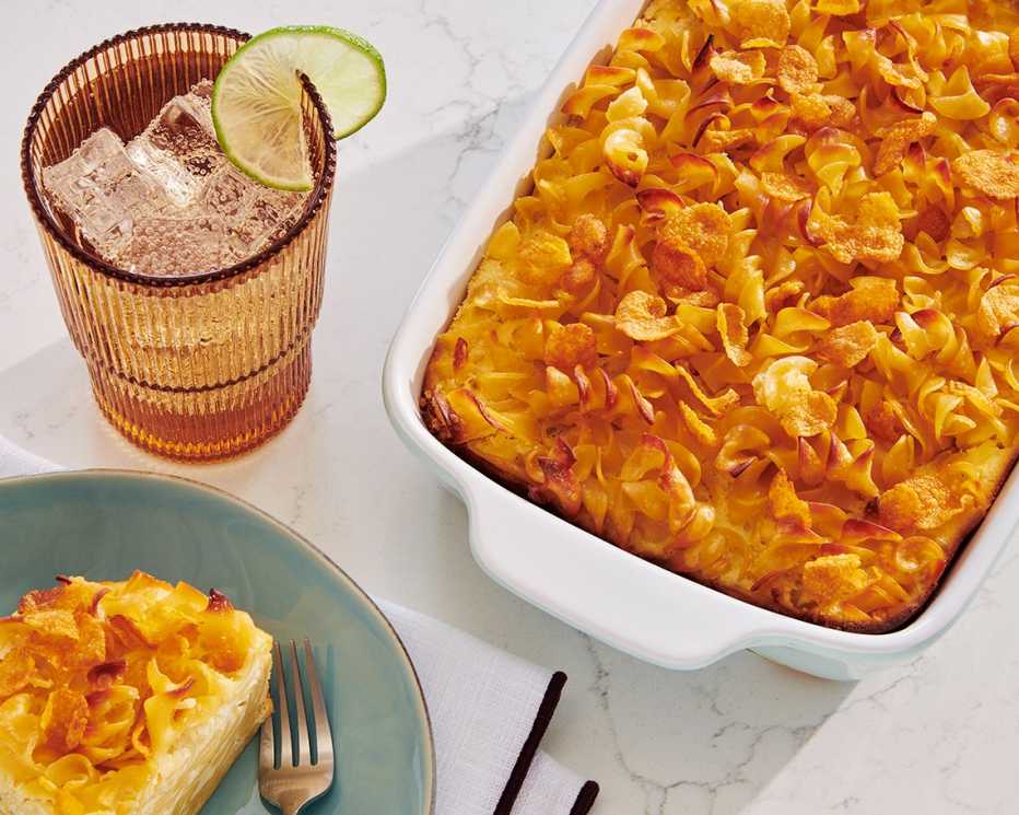 noodle pudding in a white casserole dish next to a slice on a green plate and a glass of ice tea