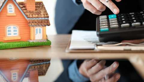 A closeup of the reflective surface of a desktop with a hand adding numbers on a calculator. A model of a house is also on the desk. A closeup of the reflective surface of a desktop with a hand adding numbers on a calculator. A model of a house is also on the desk.