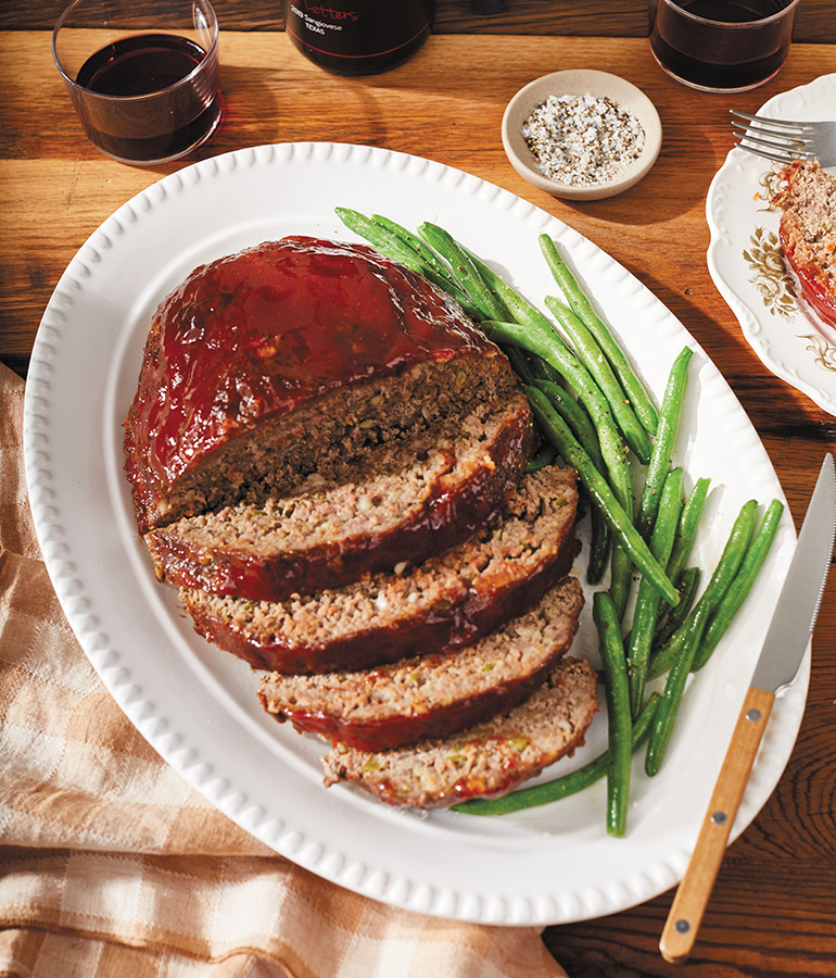 meat loaf cut into slices and green beans on plate on table; drinking glasses and some type of seasoning above plate