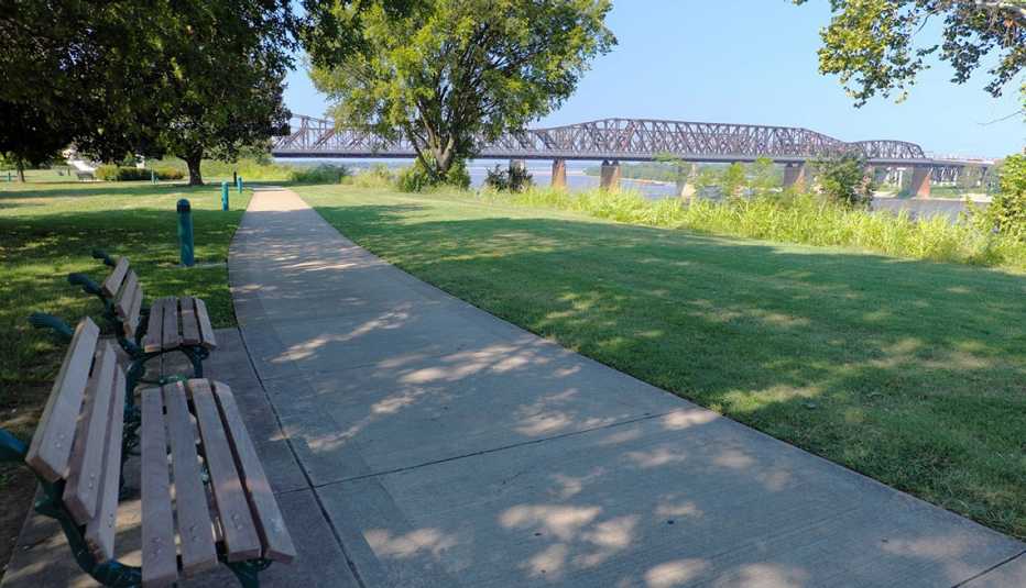 a path along a river with benches and a bridge in the background