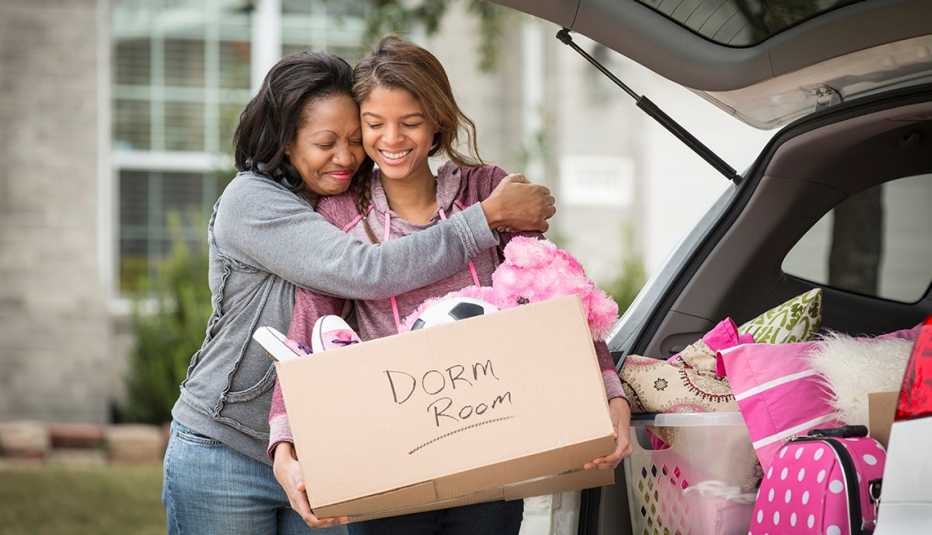 A young adult loading a box that says dorm room on it into her car as her mother hugs her