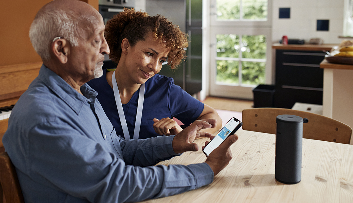 man seated in kitchen next to caretaker is learning to use a smartphone and smart speaker device