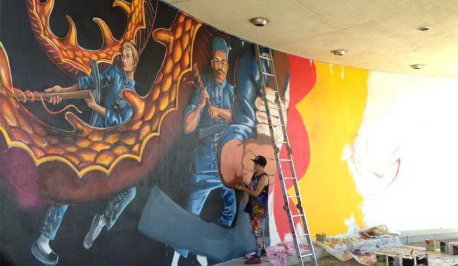 A man paints a mural showing two men fighting what looks like a dragon or octopus