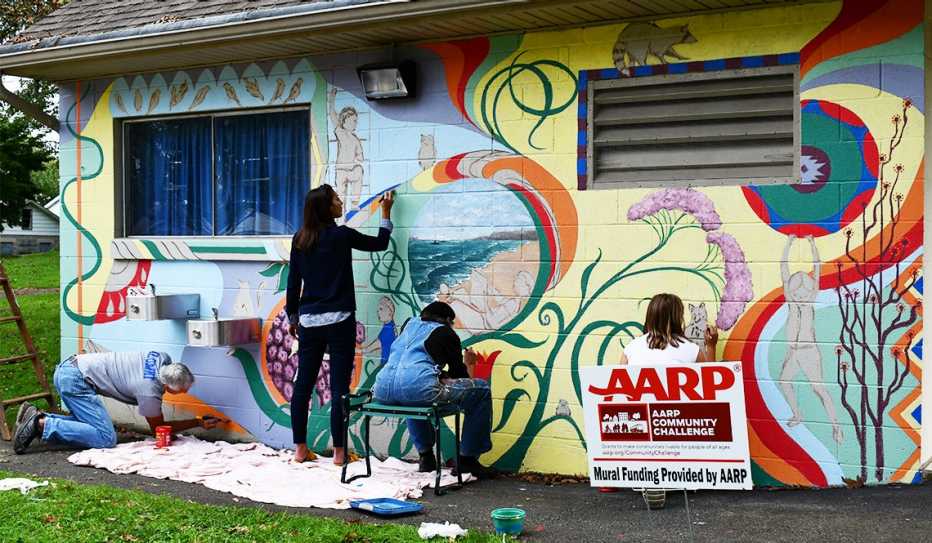 Four people paint a mural on the side of a building constructed out of cinder blocks