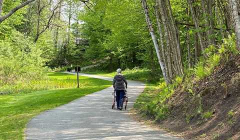An older woman pushes a wheeled walker while she travels along a multiuse recreation trail