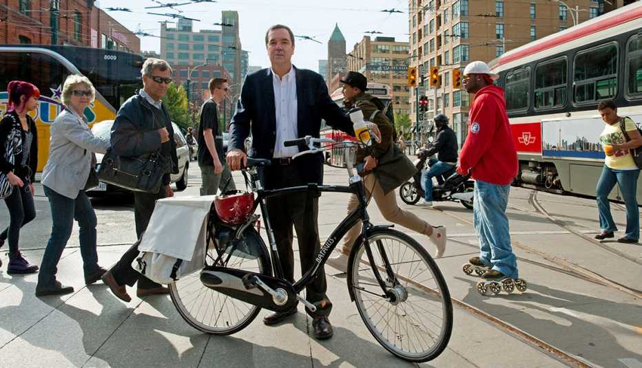 Gil Penalosa Stands In A Busy City Street With His Bike, Commuters On The Street, Buses, Livable Communities, Biking Infrastructure