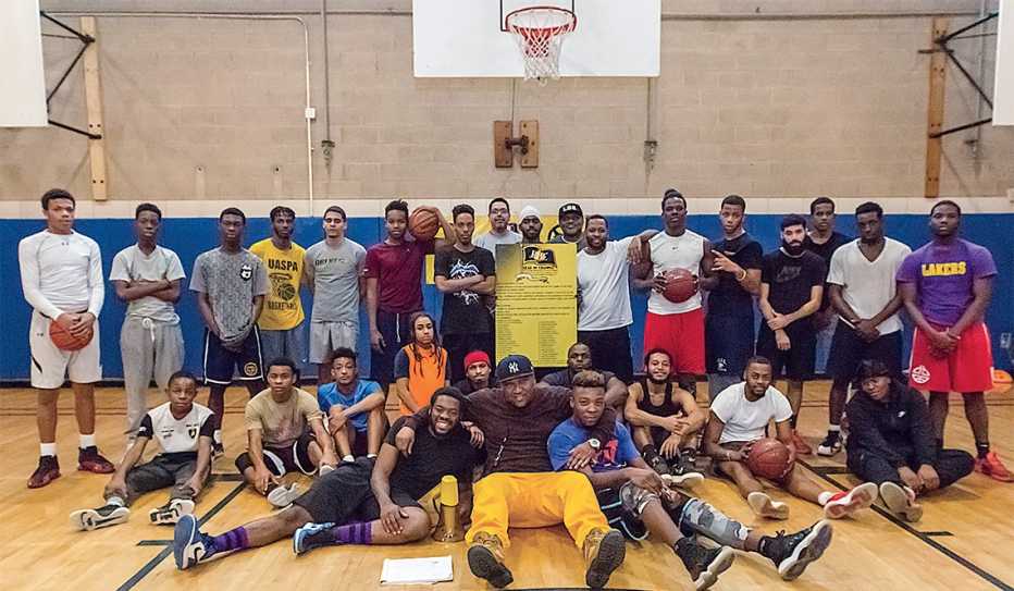 Antonio Hendrickson (seated, wearing yellow pants) poses for a basketball court group photo with young men attending a Lead by Example workshop