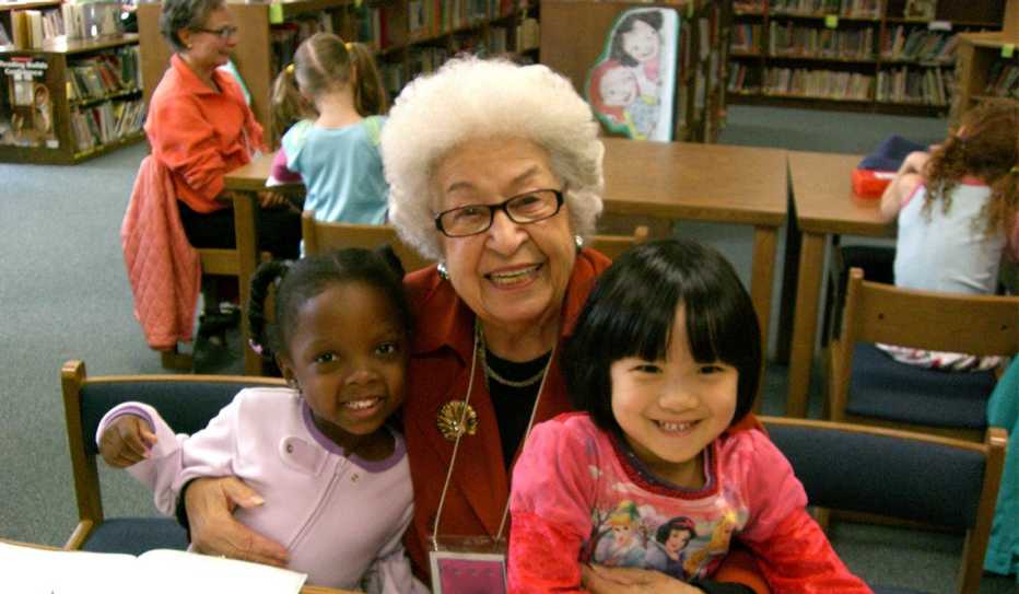 An older volunteer and two kindergartners smile during a Grandpals visit to the library An older volunteer and two kindergartners smile during a Grandpals visit to the library
