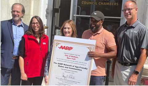 Five people (representatives from AARP New York, the New York State Department of Aging and the City of Hudson, New York), display an oversized certificate celebrating Hudson's enrollment in the AARP Network of Age-Friendly States and Communities on September 23, 2025