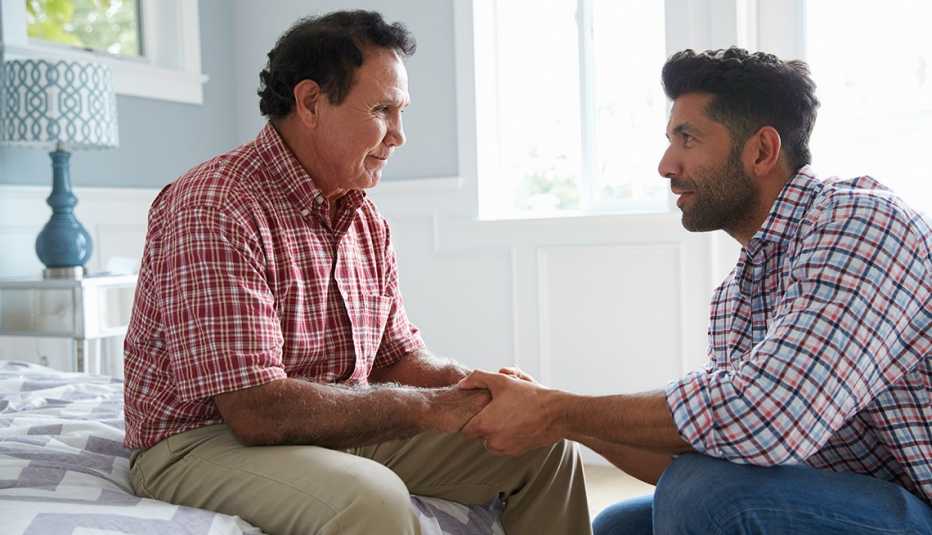 Man with dementia sits on his bed while his adult son comforts him and holds his hands