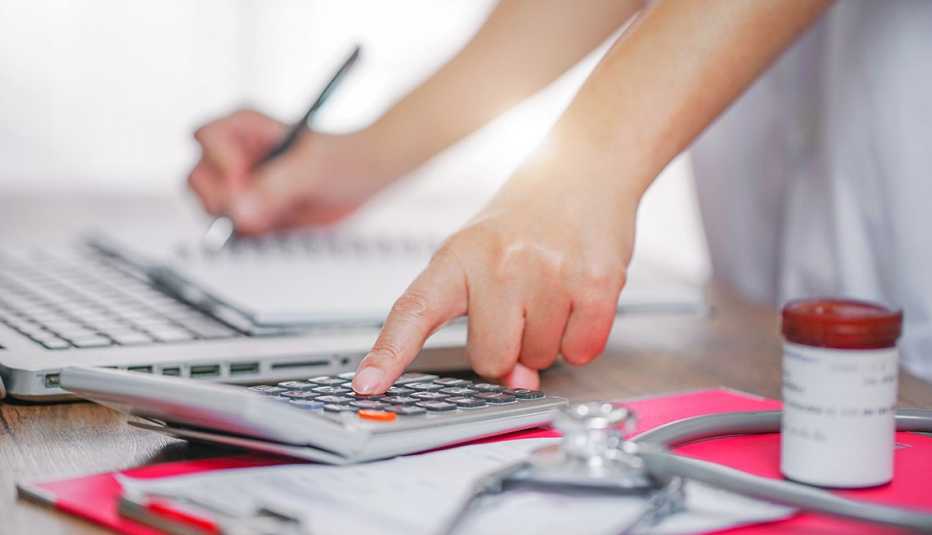 The hands of a doctor calculating cost of treatment. Stethoscope and pill bottle in foreground. 