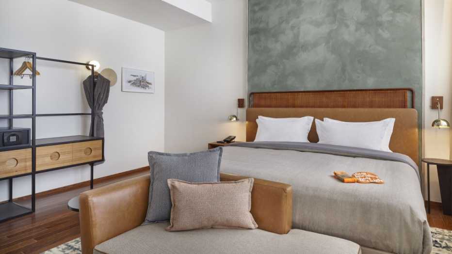 Interior of room at Hilton Canopy with bed and sofa showing in white, light brown, and white colors, wood and silver suede accents