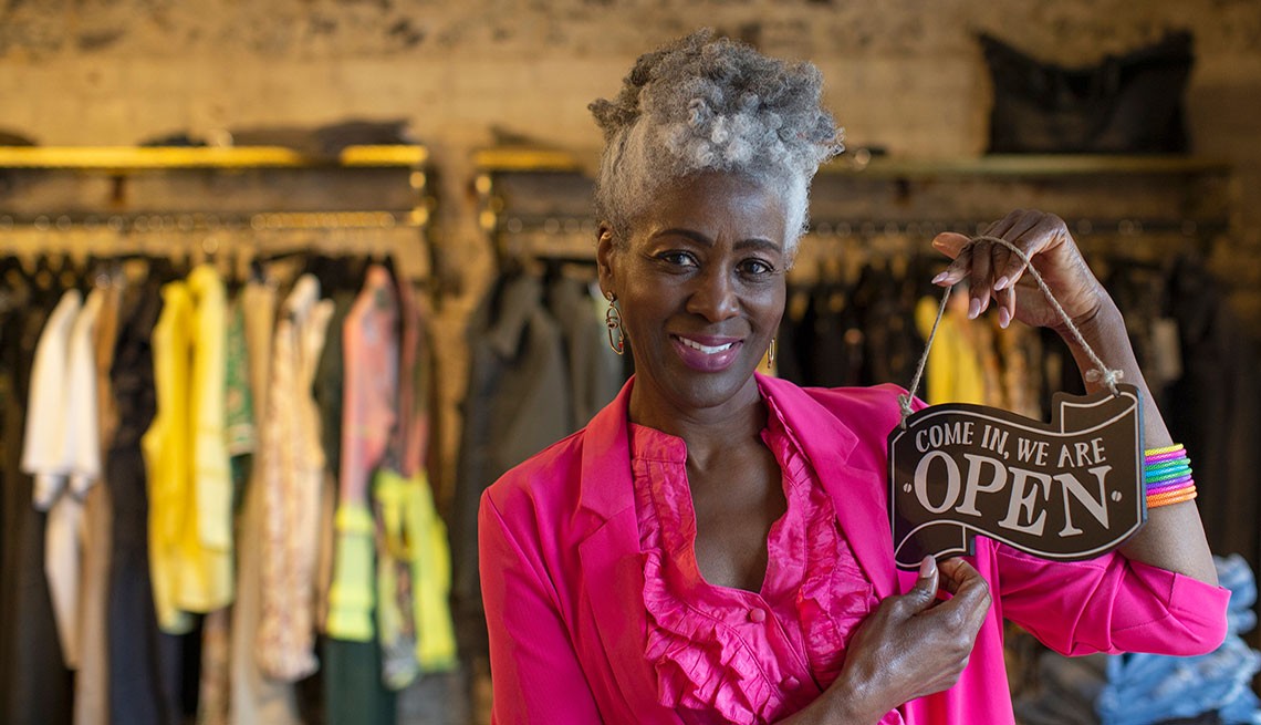 clothing store with woman in pink top and jacket holding a come in, we are open sign