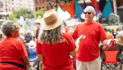 two women and one man in red t-shirts with volunteer in white lettering on back of one tee outside talking at event with other people in background