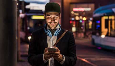 man smiling wearing black ball cap looking at cell phone with headphones around neck at night