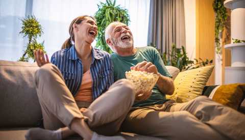 woman and man sitting on a couch, laughing, smiling, bowl of popcorn in man's hand, he is grabbing a handful, living room setting