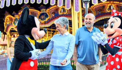 Mickey Mouse and Minnie Mouse greeting and smiling with an older man and woman in front of a lighted carousel ride