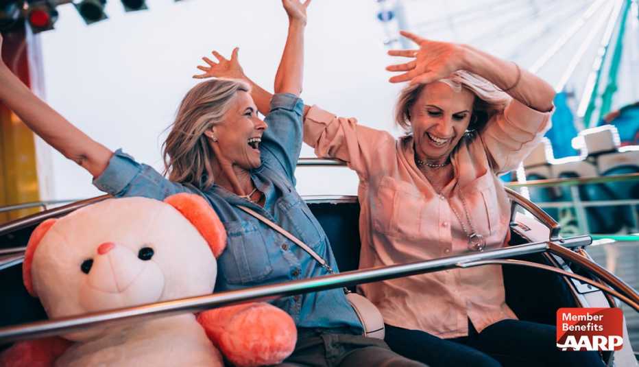 Two women riding a ride at an amusement park with a pink teddy bear in their cart and their hands in the air