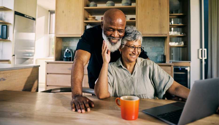 AARP member couple hugging in kitchen while looking at laptop