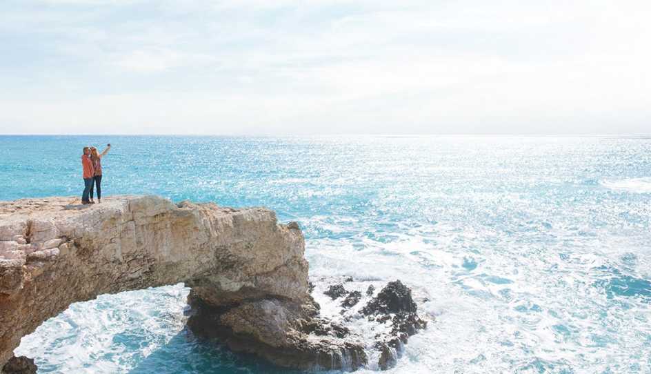 couple in long pants and long sleeves standing on a rock cliff over the ocean bright blue sky with white clouds, they are taking a selfie picture