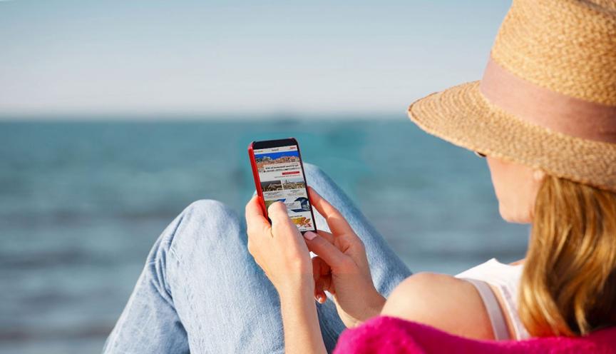 woman sitting on beach with jeans, hat, sweater, tank top, brown hat reading cellphone