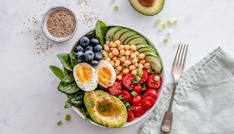 Bowl of healthy food including avocado, tomato, spinach, blueberries, hard-boiled egg, chickpeas, cucumber, green onion and chia seeds on the side with a fork to the side with a cloth napkin, light blue-gray color