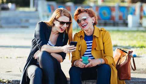 two women sitting on a street curb laughing while looking at a mobile phone