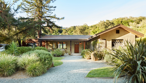tan house with rock pathway out front, surrounded by trees and shrubs
