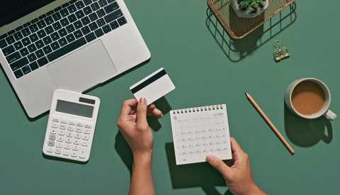 person holding a calendar and credit card at their desktop with a calculator and laptop in front of them