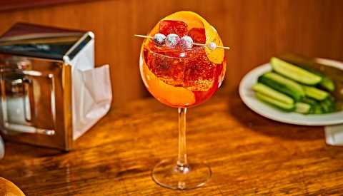 stem glass of cranberry aperol spritz on a wood table with a napkin dispenser and plate of pickles