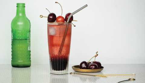 n y cherry ginger spritz in glass; cherries on right; bottle on left