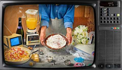 1980s TV showing someone wearing an apron holding a chocolate cream pie over a table with a bowl of spaghetti carbonara, parmesan cheese wedge, measuring spoons, an old blender with orange juice, champagne bottle, white rose bouquet, wedding figures, photo album, baby rattles and tiny old TV
