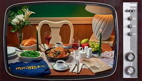 1980s TV showing skillet roast chicken on a table with green beans, glass of wine, two cups of coffee, newspaper, tall candles, bowl of fruit, flowers, salt, pepper, sugar, Michigan shirt, and a lamp
