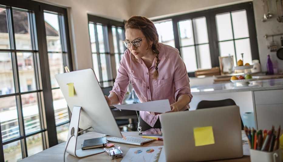woman working from home stands at dining table looking at computer screen while holding papers