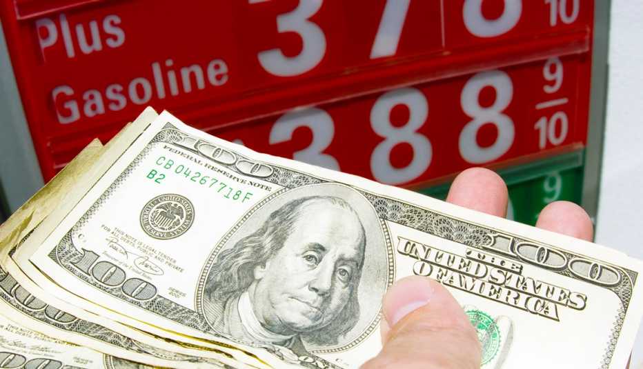 A handful of cash held in front of a high gas price sign.