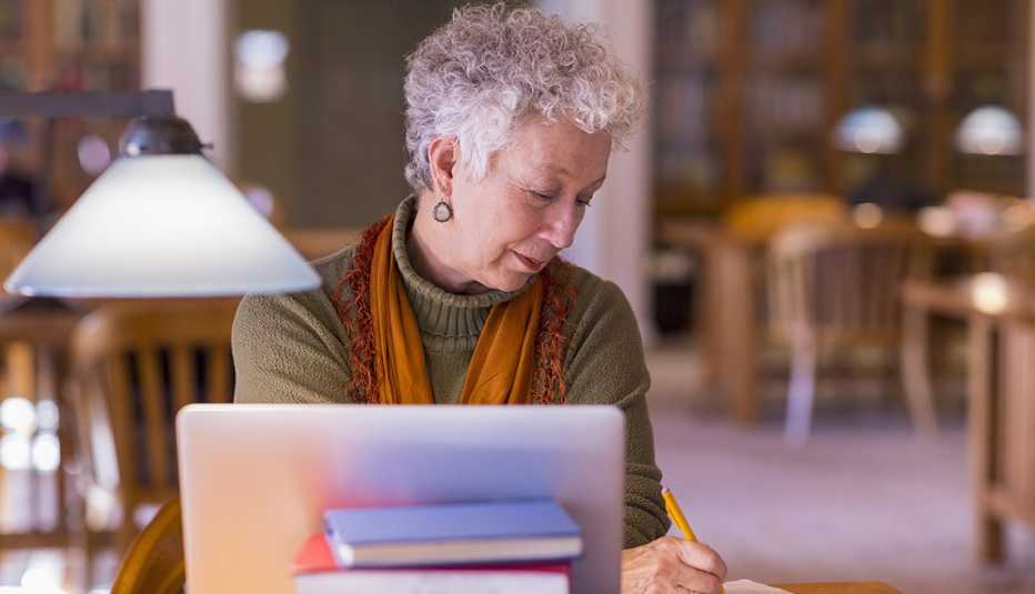  woman seated in library taking notes with laptop and stack of books in view