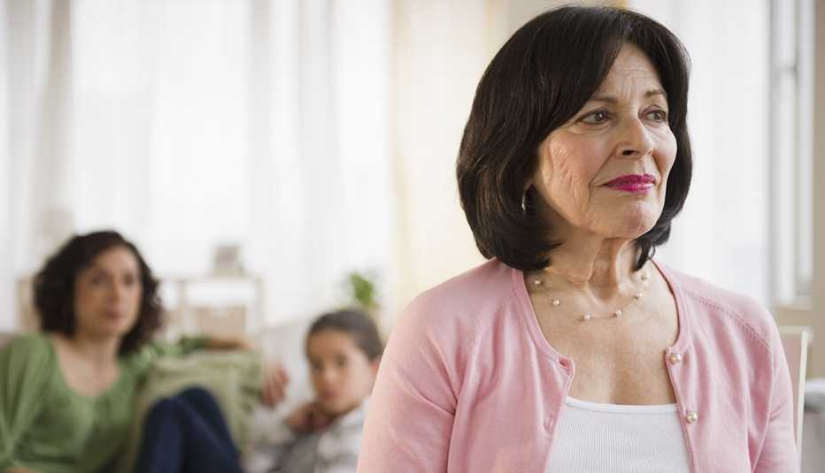 Serious women looking to her right stands in the foreground of living room, with some soft-focused family members seated in the background.