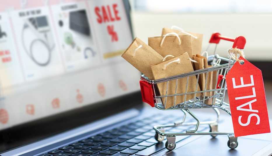 A laptop computer with an online store on screen and a miniature shopping cart full of paper gift bags, representing a cyber-shopper prepared for holiday sales deals