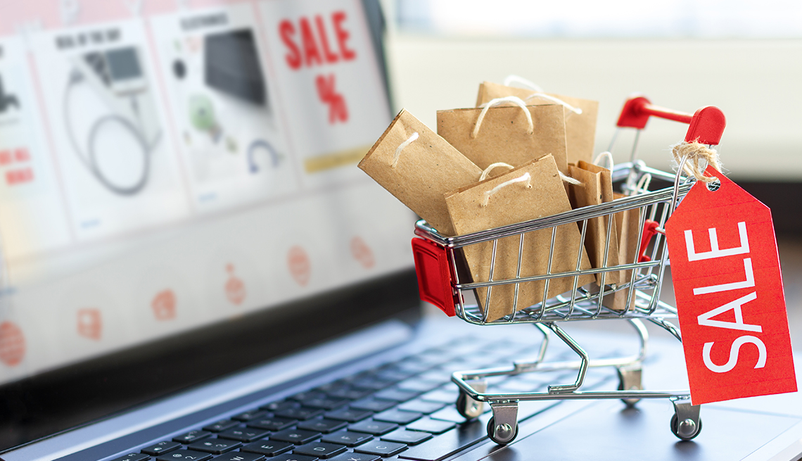 A laptop computer with an online store on screen and a miniature shopping cart full of paper gift bags, representing a cyber-shopper prepared for holiday sales deals