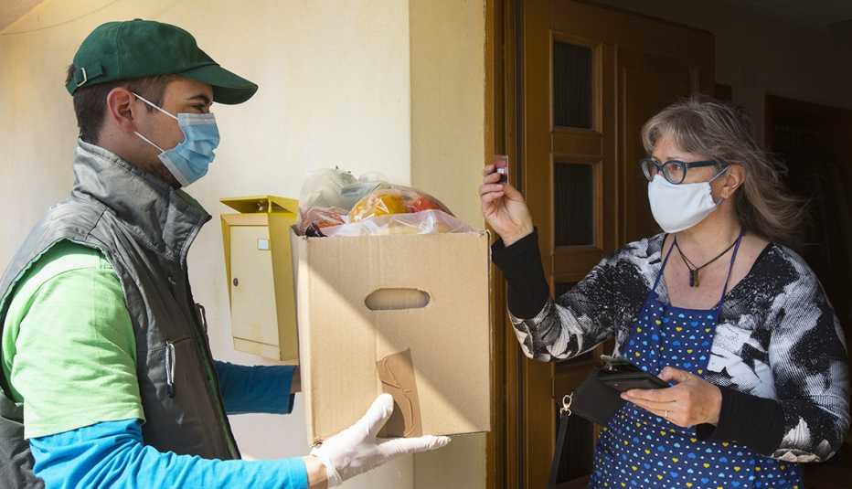 woman wearing surgical mask standing in house door way paying for home delivery of groceries with credit card