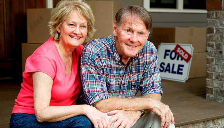 Happy senior couple sitting outside home with a red "Sold" sticker on top of the "For Sale" sign